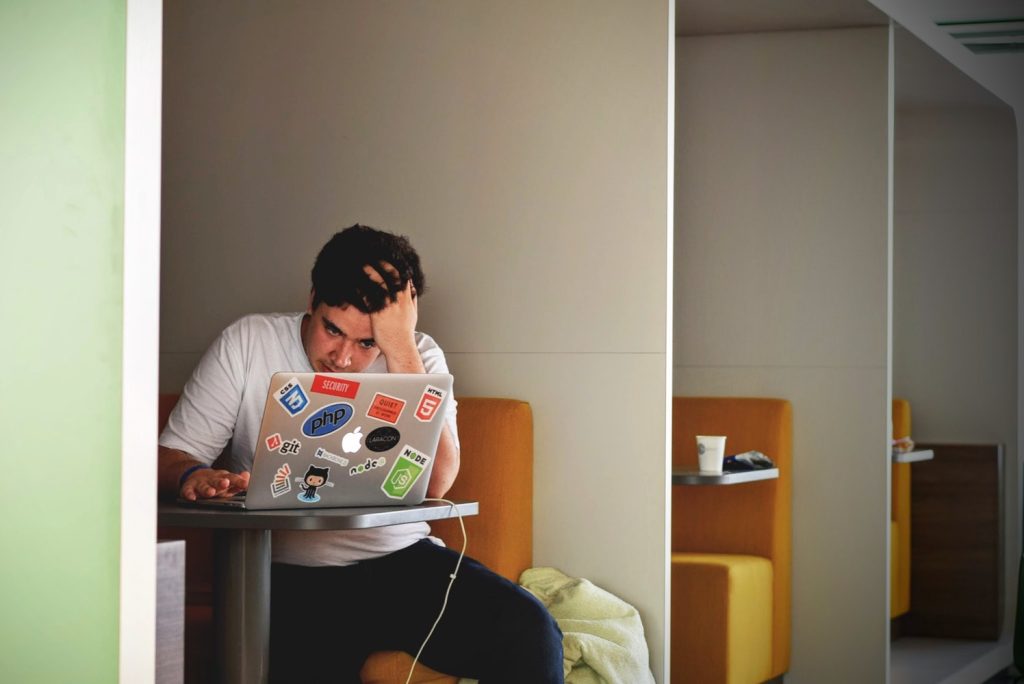 A man sits in a restaurant stall, he looks stressed and is looking at a laptop which is covered with stickers 