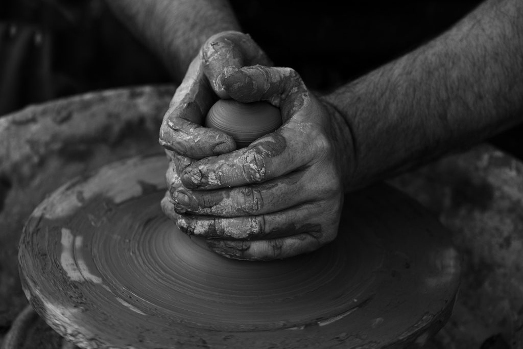 A greyscale close up photograph of someone making pottery from scratch. They are holding the wet clay in both hands while the plat spins beneath