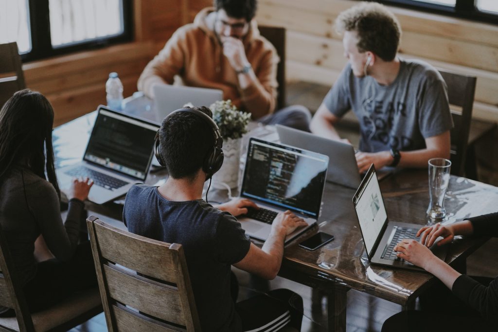 A group of five people sit around a wooden table, they are all working on laptops.