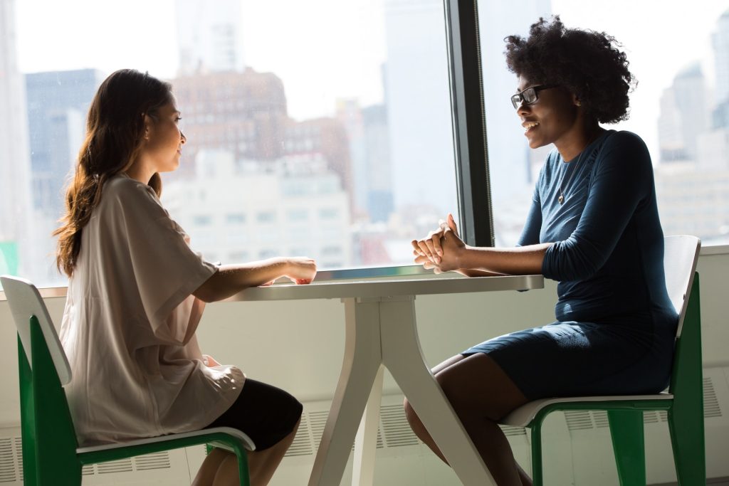 Two women sit on opposite sides of a small white circular table. The woman on the left rests both hands on the table and is listening to the lady sat across from her who has her hands clasped together and is smiling and talking 