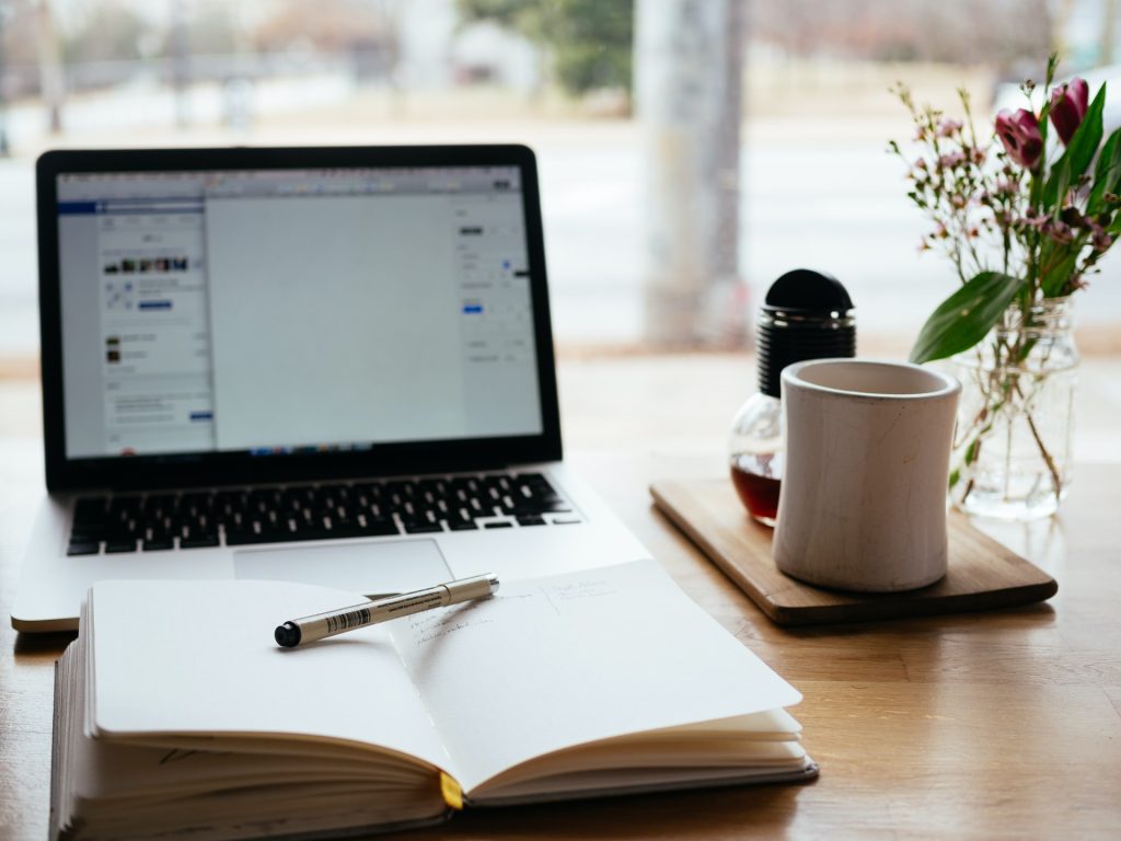 A photo of a wooden desk with a laptop sat open and an open notebook with a pen balanced on it. To the tight of there is a small glass vase of water with pink flowers and a cup standing on a small wooden board